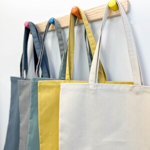 May include: Five tote bags hanging on a wooden rack with colorful hooks. The bags are in different colors: gray, blue, yellow, and white.