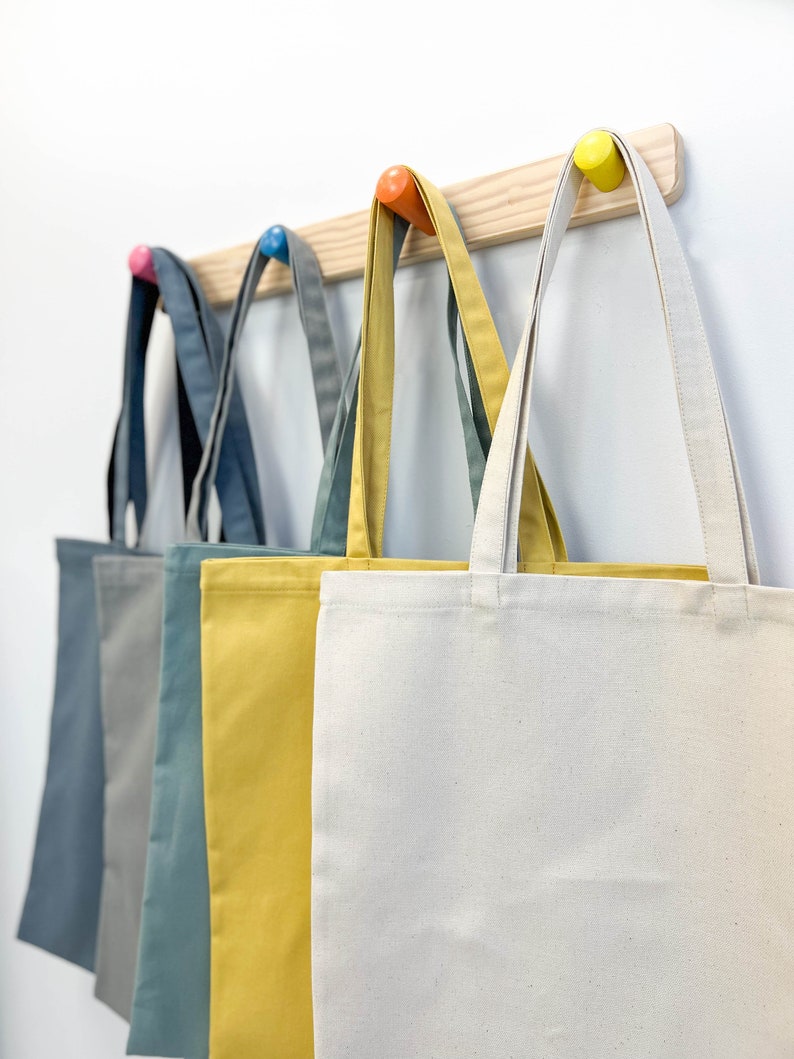 May include: Five tote bags hanging on a wooden rack with colorful hooks. The bags are in different colors: gray, blue, yellow, and white.