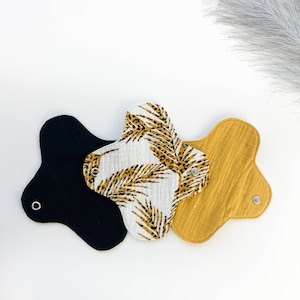 May include: Three reusable cloth menstrual pads in black, white with a gold leaf pattern, and mustard yellow. Each pad has a unique shape with snap closures and a soft, absorbent texture. The pads are designed for comfort and eco-friendliness.