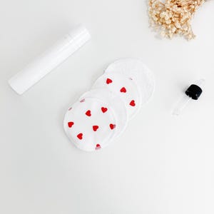 May include: White reusable facial rounds with red heart patterns are stacked near a white bottle and a glass dropper. A small bundle of dried flowers is also present. The items are arranged on a white surface, suggesting a skincare or beauty theme.