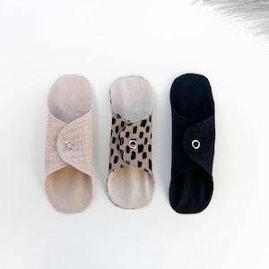 Puede incluir: Tres compresas menstruales reutilizables de tela en beige, estampado de leopardo y negro. Cada compresa está doblada y asegurada con un cierre de botón a presión plateado. Diseñadas para comodidad y ecología.