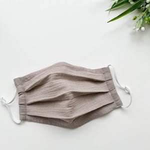 Puede incluir: Una mascarilla facial plisada de color marrón claro con presillas elásticas blancas. La mascarilla está hecha de una tela suave y texturizada. La mascarilla está diseñada para cubrir la nariz y la boca. El fondo es blanco con acentos florales verdes y blancos.