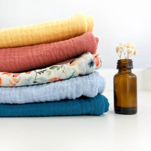 May include: A stack of five soft, colourful muslin cloths in yellow, red, blue, and white with a floral pattern. The cloths are folded and stacked on a white surface. A small brown glass bottle with a cork stopper is to the right of the cloths.