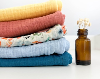 May include: A stack of five soft, colorful muslin cloths in yellow, red, blue, and white with a floral pattern. The cloths are folded and stacked on a white surface. A small brown glass bottle with a cork stopper is to the right of the cloths.