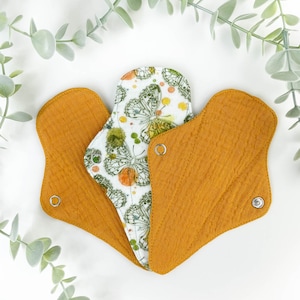 May include: Three reusable cloth menstrual pads. One pad is white with a butterfly and floral print. The other two pads are a solid mustard yellow.