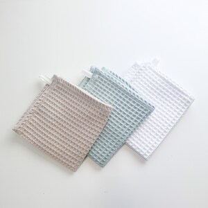 May include: Three waffle weave kitchen towels in beige, light blue, and white. The towels are folded and stacked on a white surface.