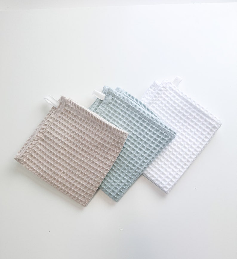 May include: Three waffle weave kitchen towels in beige, light blue, and white. The towels are folded and stacked on a white surface.