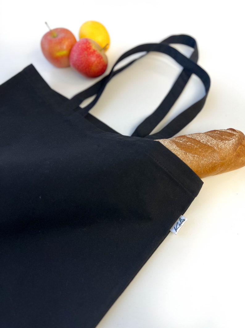 May include: A black reusable tote bag with a single baguette inside. The bag has two black straps and a white label with the word "Kila" printed on it.