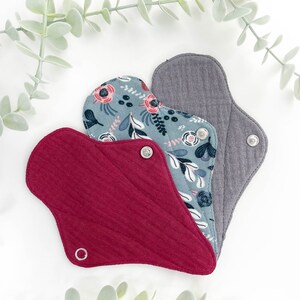 May include: Three reusable cloth menstrual pads in different colours and patterns. The pads are made of soft, absorbent fabric and have snaps for secure fastening. The pads are arranged in a triangle shape with the grey pad on top, the floral pad in the middle, and the red pad on the bottom.
