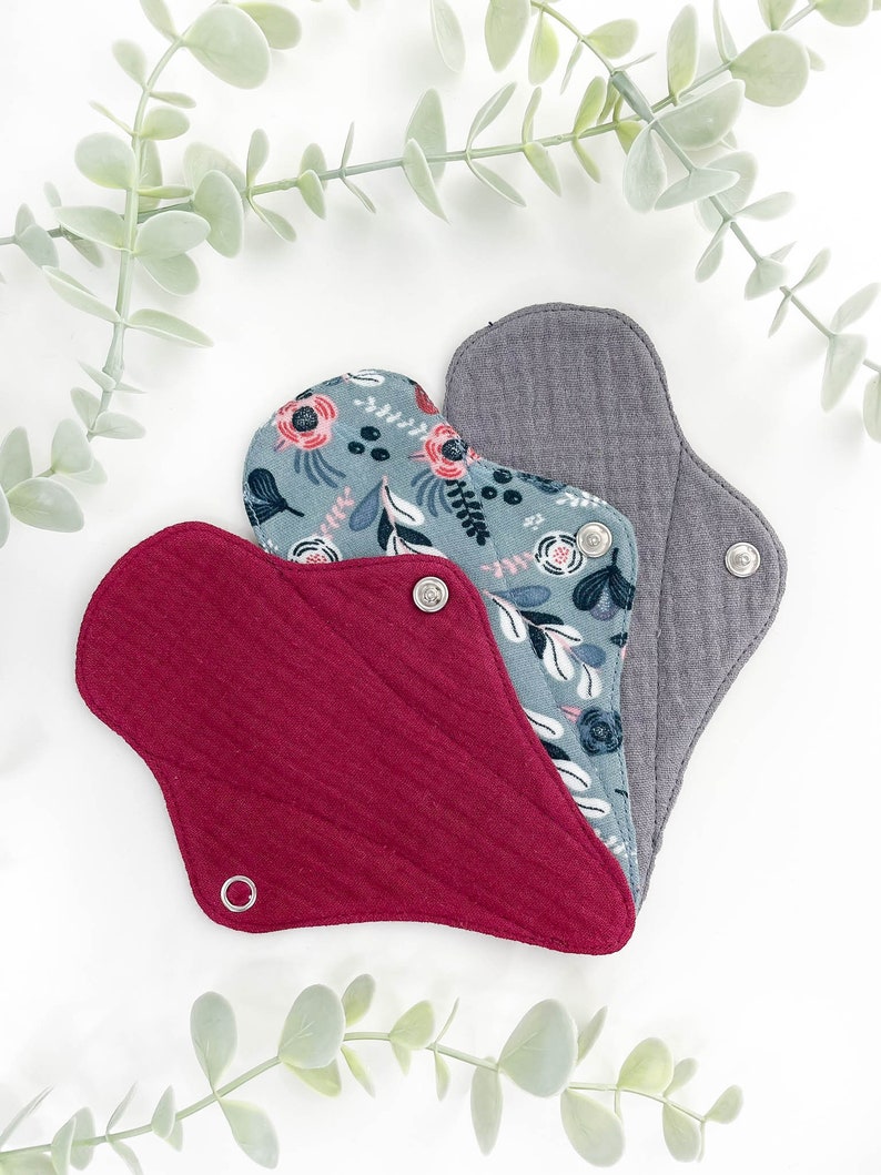 May include: Three reusable cloth menstrual pads in different colours and patterns. The pads are made of soft, absorbent fabric and have snaps for secure fastening. The pads are arranged in a triangle shape with the grey pad on top, the floral pad in the middle, and the red pad on the bottom.