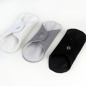 May include: Three reusable cloth menstrual pads in white, gray, and black. Each pad has a snap closure and is folded in half.