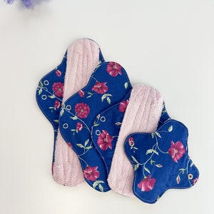 Op de afbeelding: Drie herbruikbare menstruatiedoekjes met een bloemenprint. De doekjes zijn blauw met roze bloemen en hebben een roze badstof voering.
