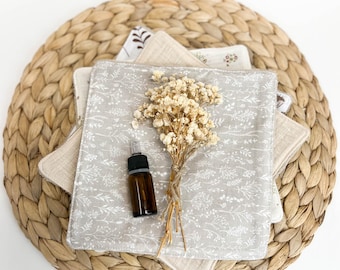 May include: A set of four beige reusable makeup remover cloths with a floral pattern, a small brown glass bottle with a dropper, and a bunch of dried white flowers on a woven straw mat.