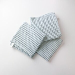 May include: Four light blue waffle weave kitchen towels stacked on a white surface.