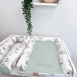 May include: A white changing pad cover featuring a jungle animal print with lions and zebras, paired with a matching cylindrical bolster pillow. A sage green waffle-textured mat with the embroidered name Matéo rests on the center of the pad.