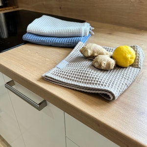 May include: Three waffle weave kitchen towels in white, blue, and beige, folded on a wooden countertop. A lemon and ginger root are on the beige towel.