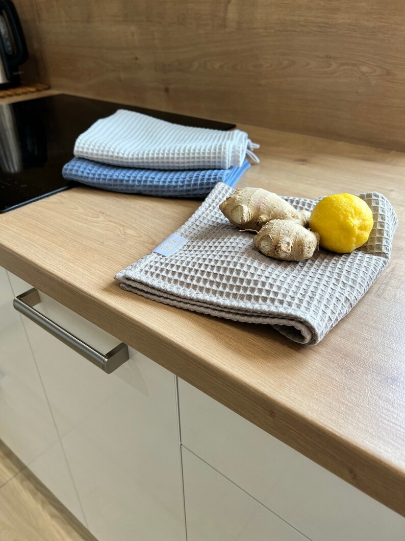 May include: Three waffle weave kitchen towels in white, blue, and beige, folded on a wooden countertop. A lemon and ginger root are on the beige towel.