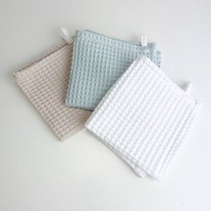 May include: Three waffle weave kitchen towels in beige, light blue, and white. The towels are folded and arranged on a white surface.