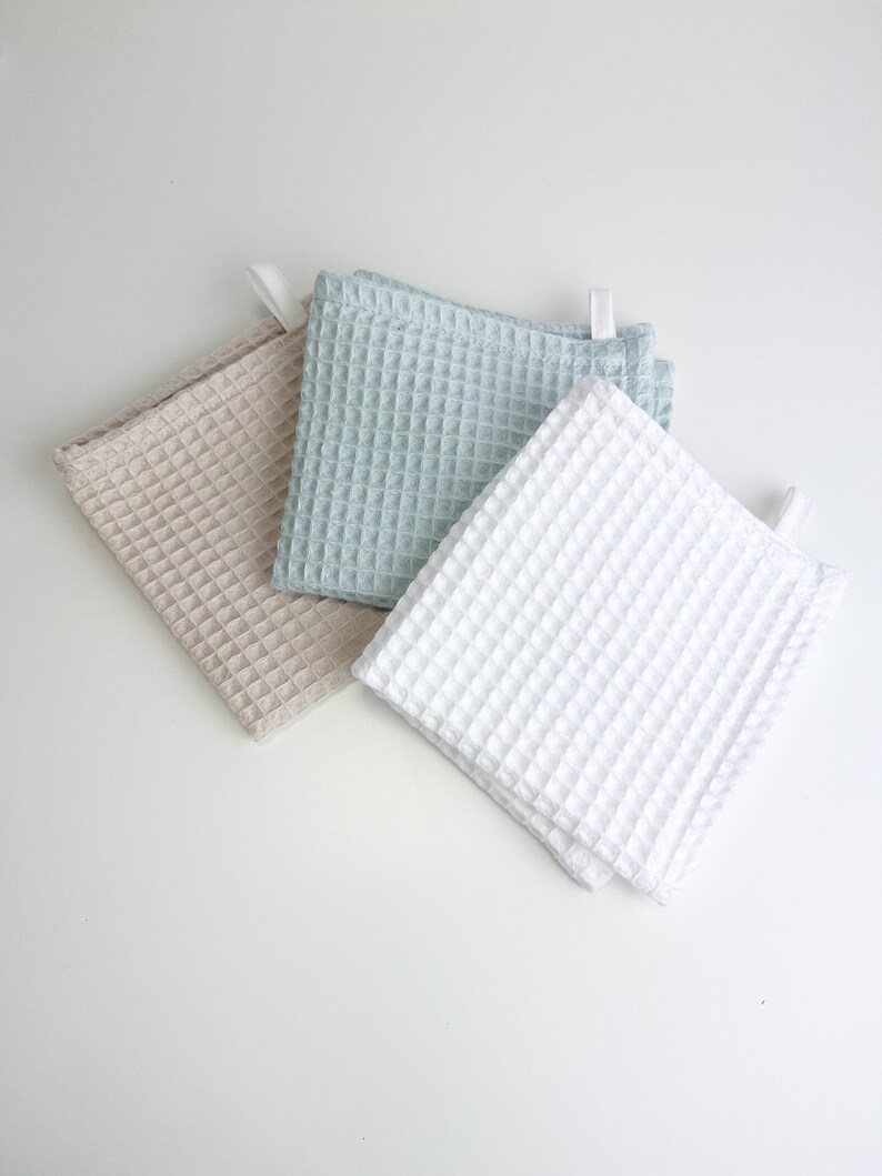 May include: Three waffle weave kitchen towels in beige, light blue, and white. The towels are folded and arranged on a white surface.
