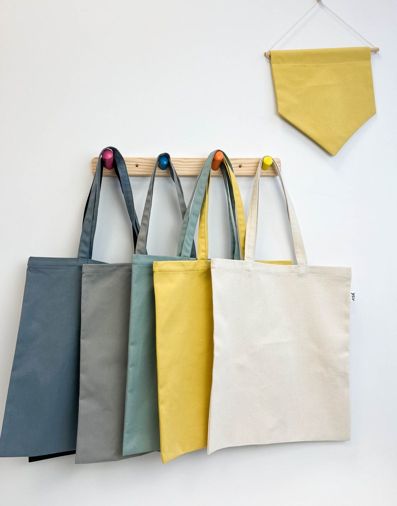 May include: Five tote bags hanging on a wooden rack with colorful knobs. The bags are in shades of blue, gray, green, yellow, and white.