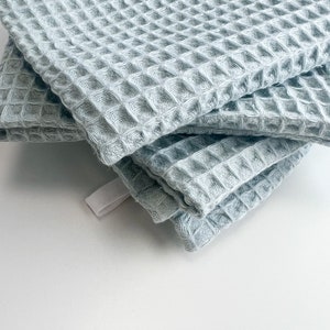 May include: A stack of four light blue waffle weave kitchen towels. The towels are folded and have a textured waffle pattern.