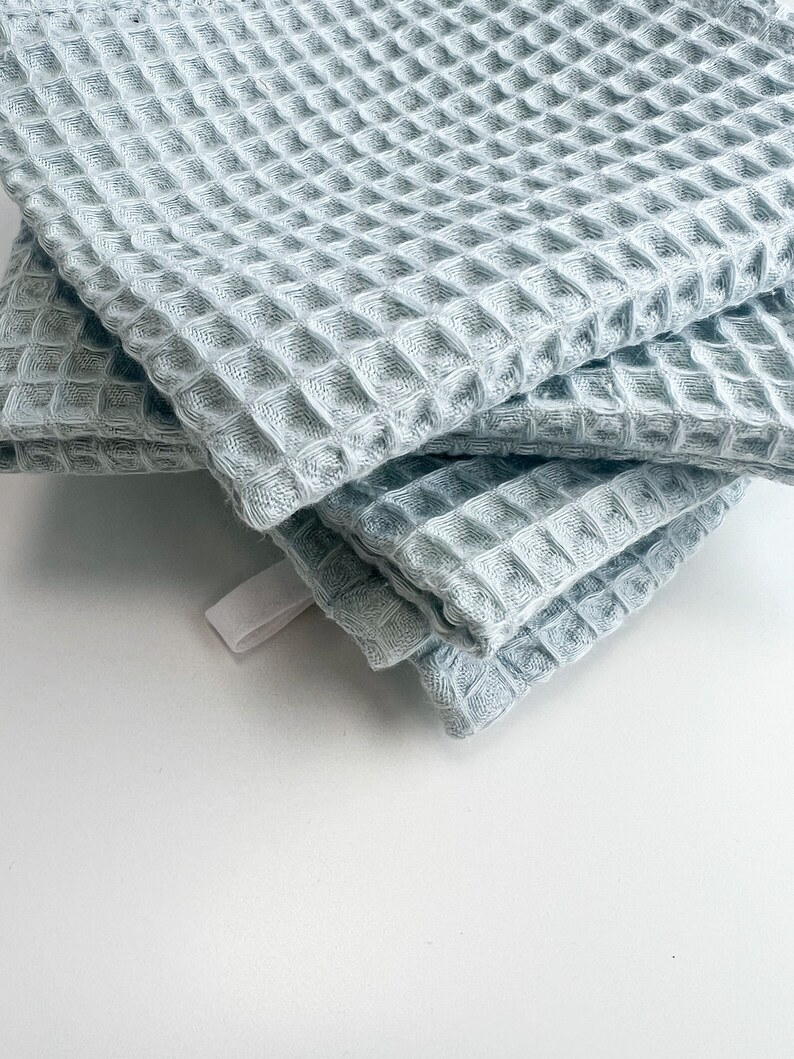 May include: A stack of four light blue waffle weave kitchen towels. The towels are folded and have a textured waffle pattern.