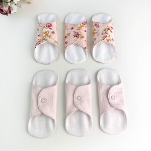 May include: Six reusable cloth menstrual pads. Three pads have a pink floral pattern, and three are solid light pink. The pads are folded and secured with a snap closure, with a white absorbent interior.
