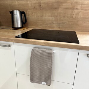 May include: A gray waffle weave kitchen towel hangs on a white cabinet door handle. The towel is folded in half and has a small white tag with the word "Oeko-Tex" on it.
