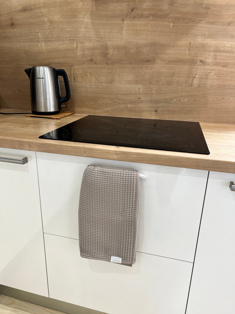 May include: A gray waffle weave kitchen towel hangs on a white cabinet door handle. The towel is folded in half and has a small white tag with the word "Oeko-Tex" on it.