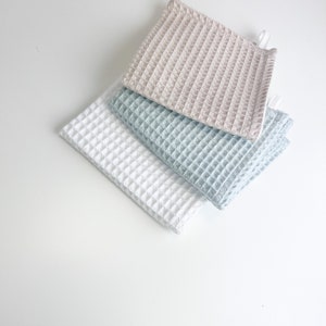May include: Three waffle weave kitchen towels in beige, light blue, and white. The towels are folded and stacked on top of each other.