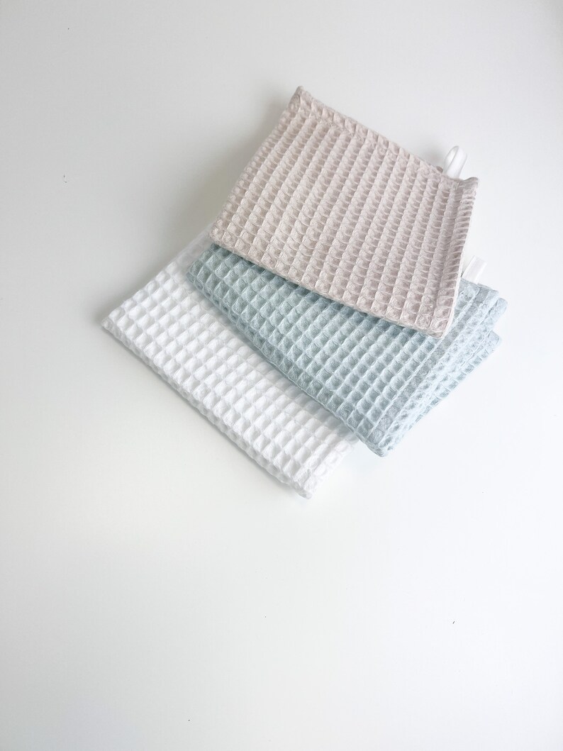 May include: Three waffle weave kitchen towels in beige, light blue, and white. The towels are folded and stacked on top of each other.