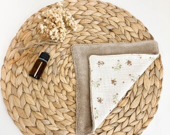 May include: A brown essential oil bottle and two baby bibs rest on a woven placemat. One bib is solid brown, and the other is white with a floral pattern. Dried flowers are placed near the bottle.