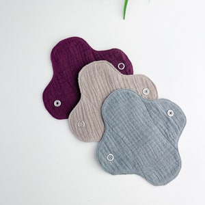 Può includere: Tre assorbenti mestruali in tessuto riutilizzabili in diversi colori: bordeaux, beige e grigio. Gli assorbenti sono realizzati in tessuto morbido e assorbente e hanno un design unico a forma di fiore.