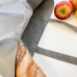 May include: A gray canvas tote bag with a long loaf of bread inside. The bag has two straps and is open at the top. There are three red and yellow apples on a white surface next to the bag.