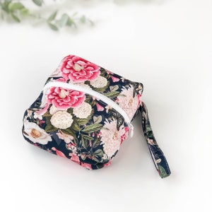 Puede incluir: Una pequeña bolsa cuadrada con cremallera de estampado floral con una correa negra. La bolsa tiene un fondo azul oscuro con flores rosas y blancas.