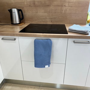Puede incluir: Una cocina blanca con una tetera de acero inoxidable sobre una encimera de madera. La encimera tiene una placa de cocci&oacute;n el&eacute;ctrica negra y un pa&ntilde;o de cocina azul de tejido de gofre colgado de una manija de armario.