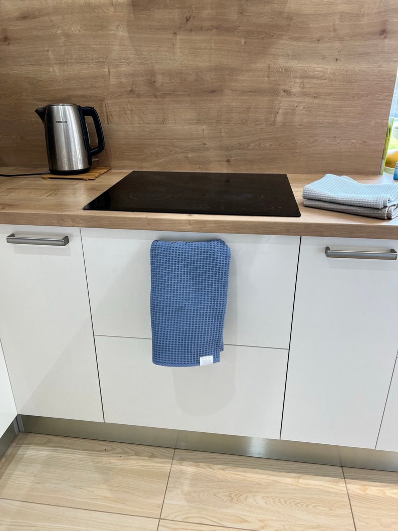 Puede incluir: Una cocina blanca con una tetera de acero inoxidable sobre una encimera de madera. La encimera tiene una placa de cocci&oacute;n el&eacute;ctrica negra y un pa&ntilde;o de cocina azul de tejido de gofre colgado de una manija de armario.