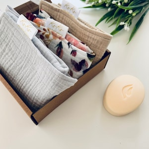 May include: A gift box filled with three cotton muslin cloths, one gray, one beige, and one with a floral print. The cloths have the text "Koko Zero Waste Products" printed on them. A bar of soap with a leaf design is sitting on a white surface next to the box.