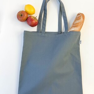 May include: A gray canvas tote bag with two handles. The bag is empty, but there are two red apples and a yellow lemon on the white surface next to it. A loaf of bread is sticking out of the top of the bag.