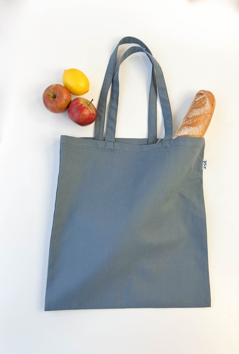 May include: A gray canvas tote bag with two handles. The bag is empty, but there are two red apples and a yellow lemon on the white surface next to it. A loaf of bread is sticking out of the top of the bag.