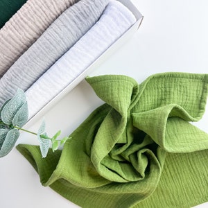 Peut inclure: Un ensemble de cinq linges en mousseline dans une bo&icirc;te blanche. Les linges sont dans des tons de vert, beige, gris et blanc. Un seul linge en mousseline vert est pli&eacute; et plac&eacute; sur une surface blanche.