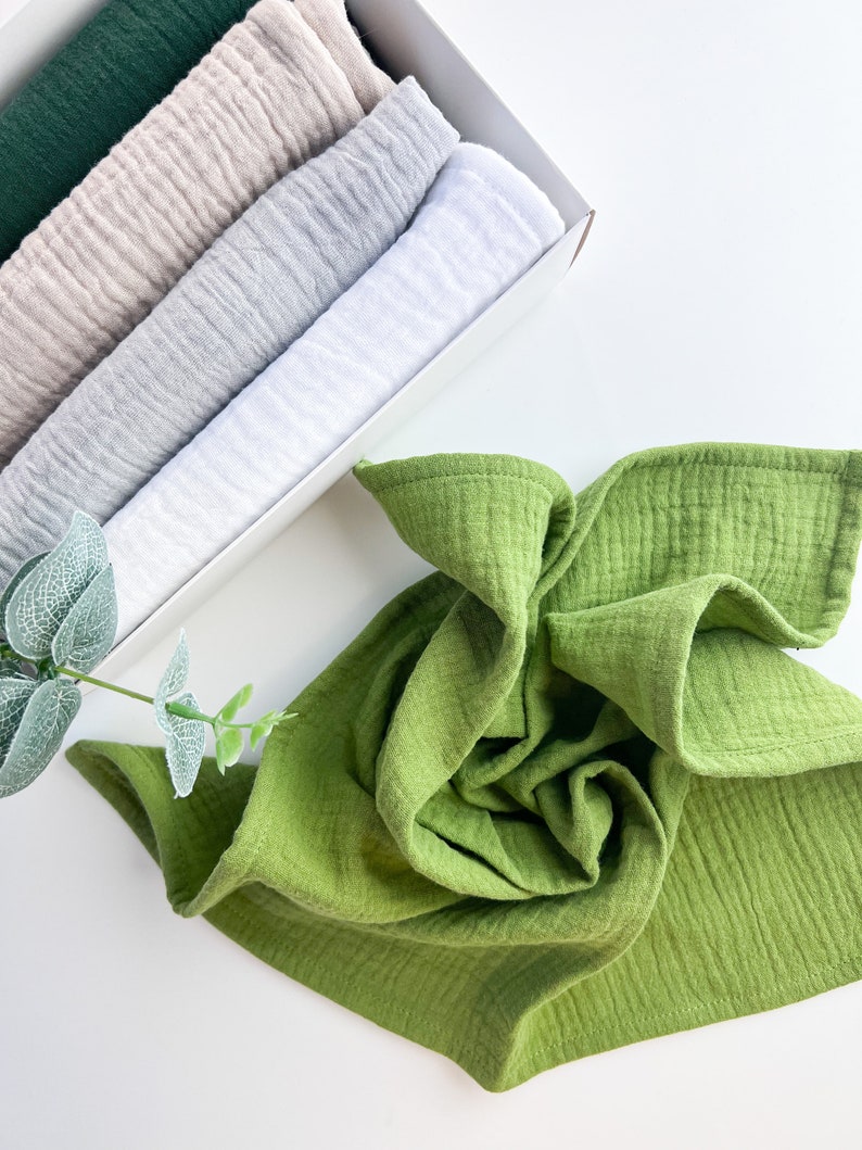 Peut inclure: Un ensemble de cinq linges en mousseline dans une bo&icirc;te blanche. Les linges sont dans des tons de vert, beige, gris et blanc. Un seul linge en mousseline vert est pli&eacute; et plac&eacute; sur une surface blanche.