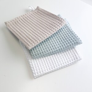 May include: Three waffle weave kitchen towels in beige, light blue, and white. The towels are stacked on top of each other.