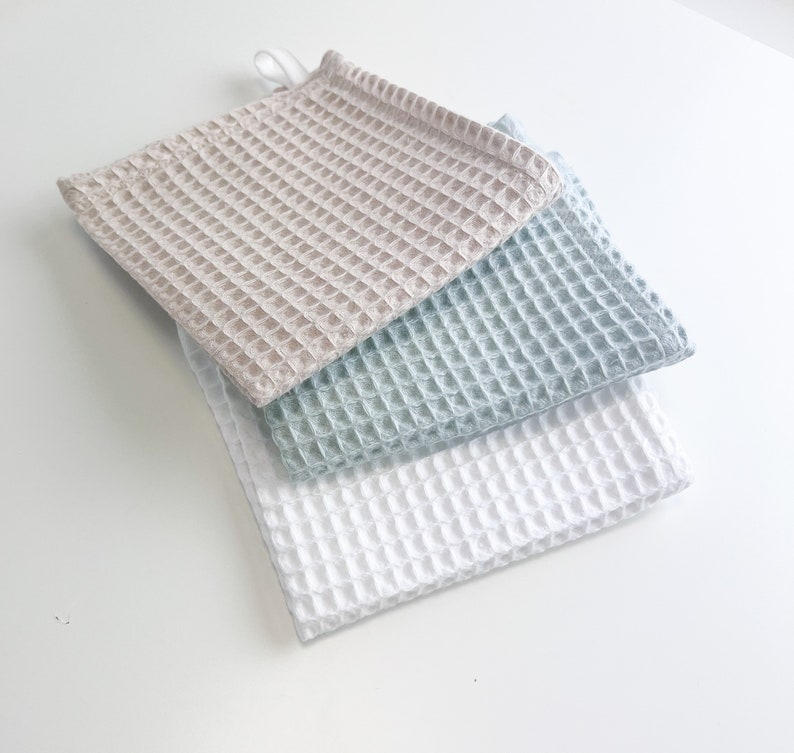 May include: Three waffle weave kitchen towels in beige, light blue, and white. The towels are stacked on top of each other.