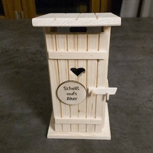 Könnte beinhalten: Ein kleines Modell eines Holz-Plumpsklos, aus hellem Holz gefertigt. Die Tür hat einen herzförmigen Ausschnitt und ein kleines Schild mit der Aufschrift "Scheiß aufs Alter". Das Dach ist mit Lamellen versehen, und die Tür hat einen kleinen Riegel.