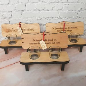 Könnte beinhalten: Holz-Schnapsglashalter in Form von Bänken. Jede Bank hat ein kleines Glas und ein Holzschild mit deutschem Text. Die Bänke sind aus hellem Holz mit schwarzen Akzenten und mit roter Schnur verziert.