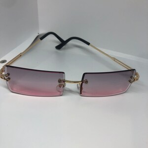 Pode incluir: Óculos de sol com armação dourada e lentes com degradê rosa. Os óculos têm uma armação retangular e uma ponte nasal fina e preta.