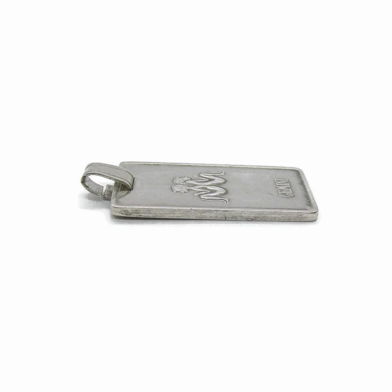 Puede incluir: Pendiente de plata con forma rectangular y un logotipo de dos serpientes entrelazadas con la palabra "JUNO" grabada en la parte inferior.