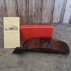 May include: A vintage Electro-Static Cleaner in a brown and black plastic case with a brush. The cleaner is designed to remove dust and lint from surfaces using static electricity. The box is red and has the words "New Electro-Static Cleaner" printed on it.