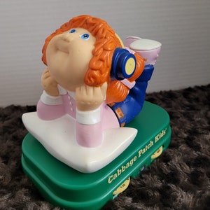 May include: A Cabbage Patch Kids toy featuring a red-haired girl wearing headphones and a pink dress. The toy sits on a green base with a white pillow and the words "Cabbage Patch Kids" printed on the side.